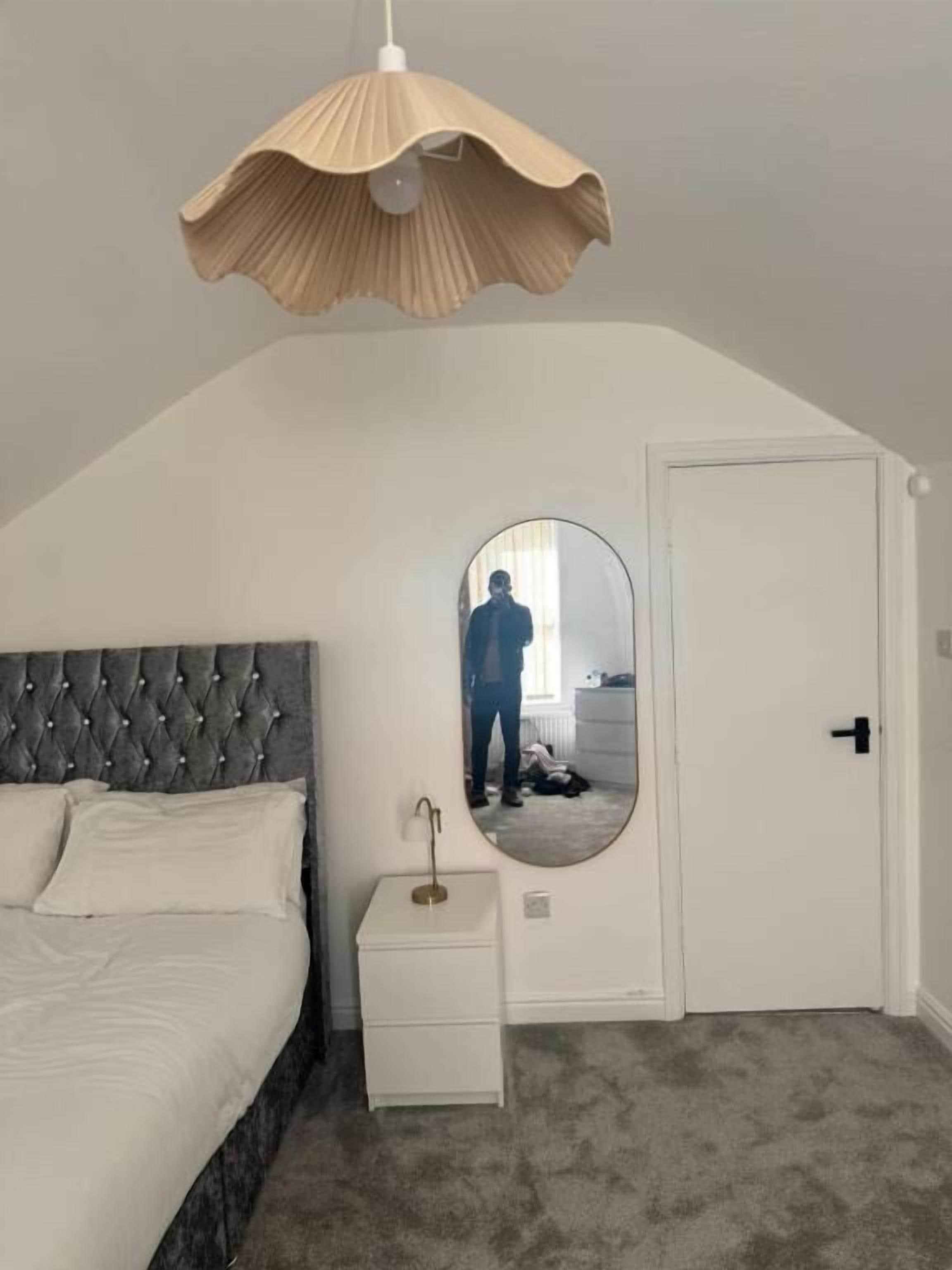 Slaapkamer met een gestoffeerd hoofdbord, nachtkastje met een lamp, grote spiegel en een deur.