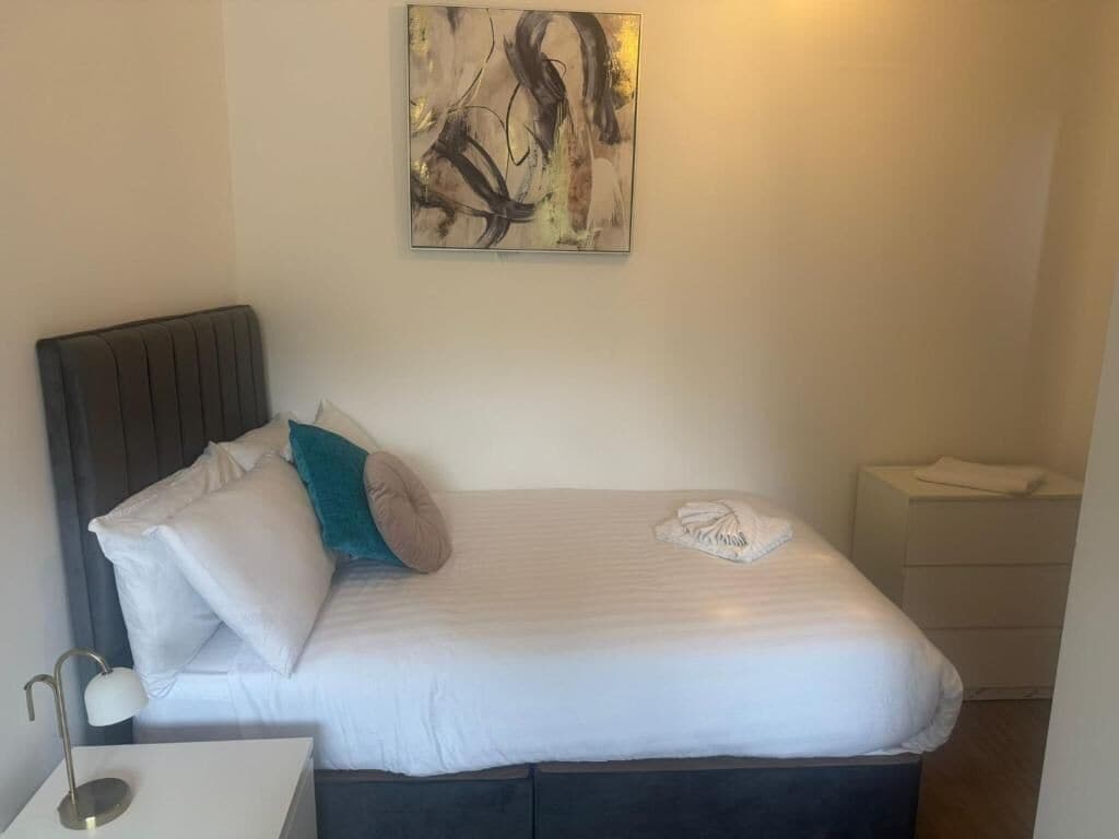 Una cama bien hecha con ropa de cama blanca y algunos almohadones, una mesita de noche con lámpara, y un cuadro abstracto colgado en la pared.