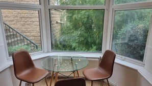 Una habitación con una mesa de vidrio y dos sillas, ventanas amplias con vistas a la vegetación, y una pared de ladrillo.
