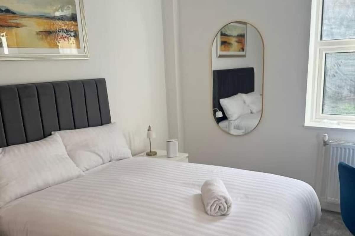 Un lit bien fait, avec une serviette pliée, une table de chevet avec une lampe, un miroir fixé au mur et un tableau au-dessus du lit.