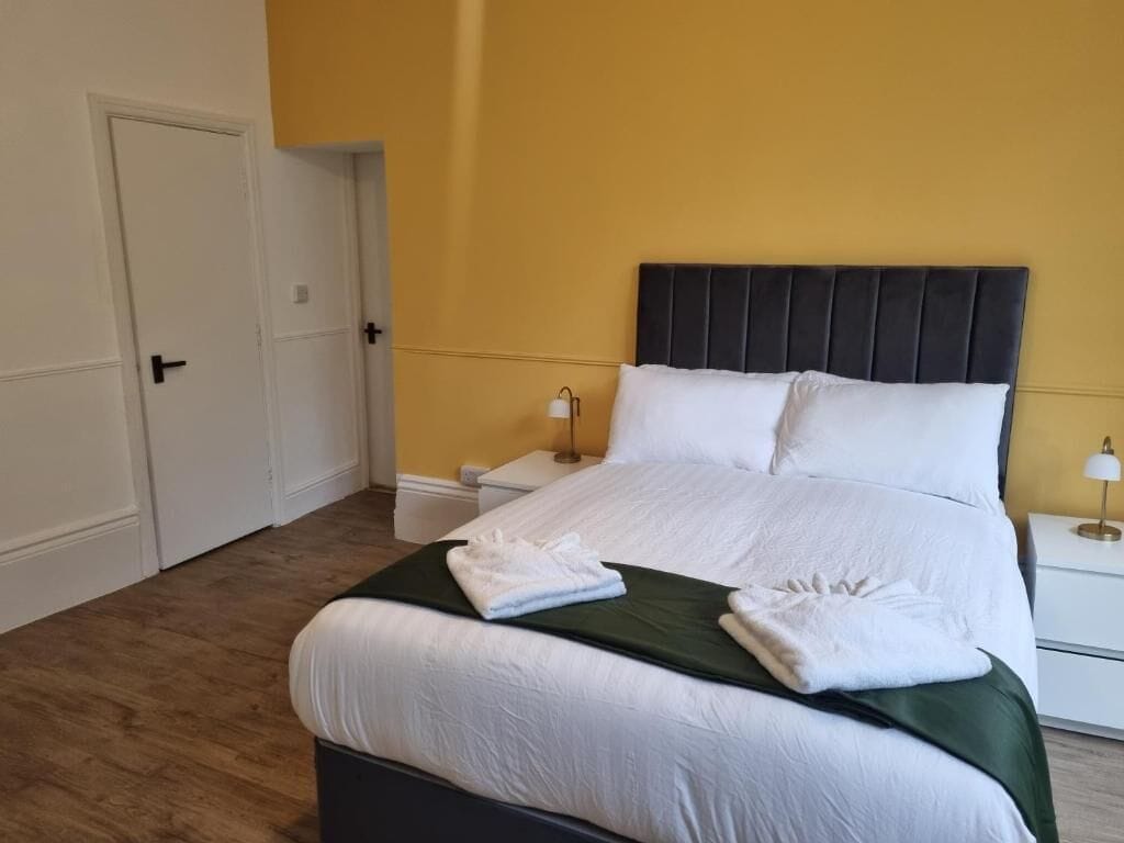 Una cama bien hecha con sábanas blancas y un edredón verde, dos mesitas de noche con lámparas y una puerta a la izquierda.