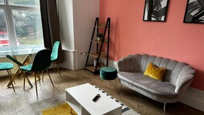 Una sala moderna con una mesa de vidrio, sillas color turquesa, una mesa de centro blanca, un sofá gris con un cojín amarillo y una escalera apoyada contra la pared.