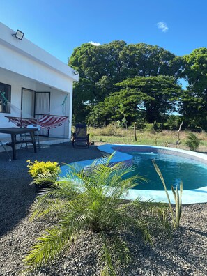En pool med kakelbeklädd kant, omgiven av grus och små växter. Utanför en byggnad med vit fasad och en dörr finns en hängmatta och ett bord med stolar.