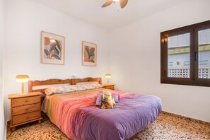 Un dormitorio con una cama, dos mesitas de noche, un ventilador de techo, dos cuadros enmarcados y una ventana con vistas.