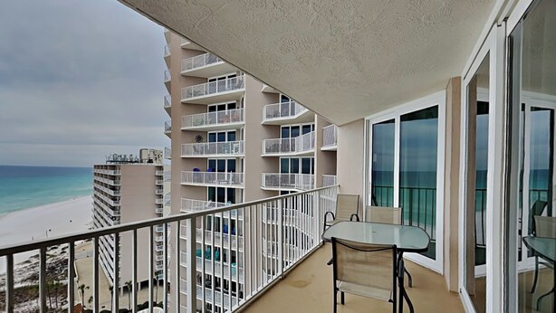 Ein Balkon mit Glastisch und -stühlen, der Blick auf einen Strand und ein Schwimmbad freigibt.