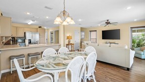 A modern kitchen with white cabinets, a stainless steel refrigerator, and a dining area with a glass table and white chairs. - The Turtle's Nest (Port St. Joe)