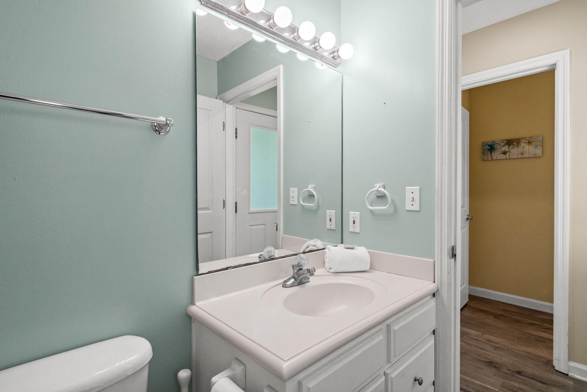 Un baño con un lavamanos blanco, un espejo con iluminación, un perchero para toallas y una puerta que da a otra habitación.