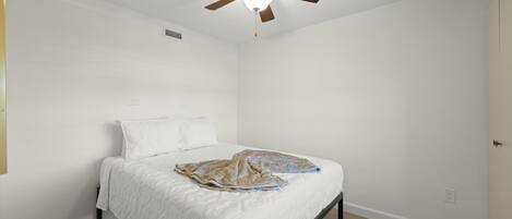 Un dormitorio con una cama, paredes blancas, un ventilador de techo y suelo de madera.