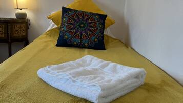 Una cama con una manta amarilla y una toalla blanca doblada encima. Hay un cojín decorativo con un estampado colorido.