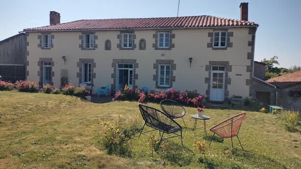 A large, two-story building with a tiled roof, multiple windows, and a small garden area with chairs and flowers. - Au pré du Puy (Chanverrie)