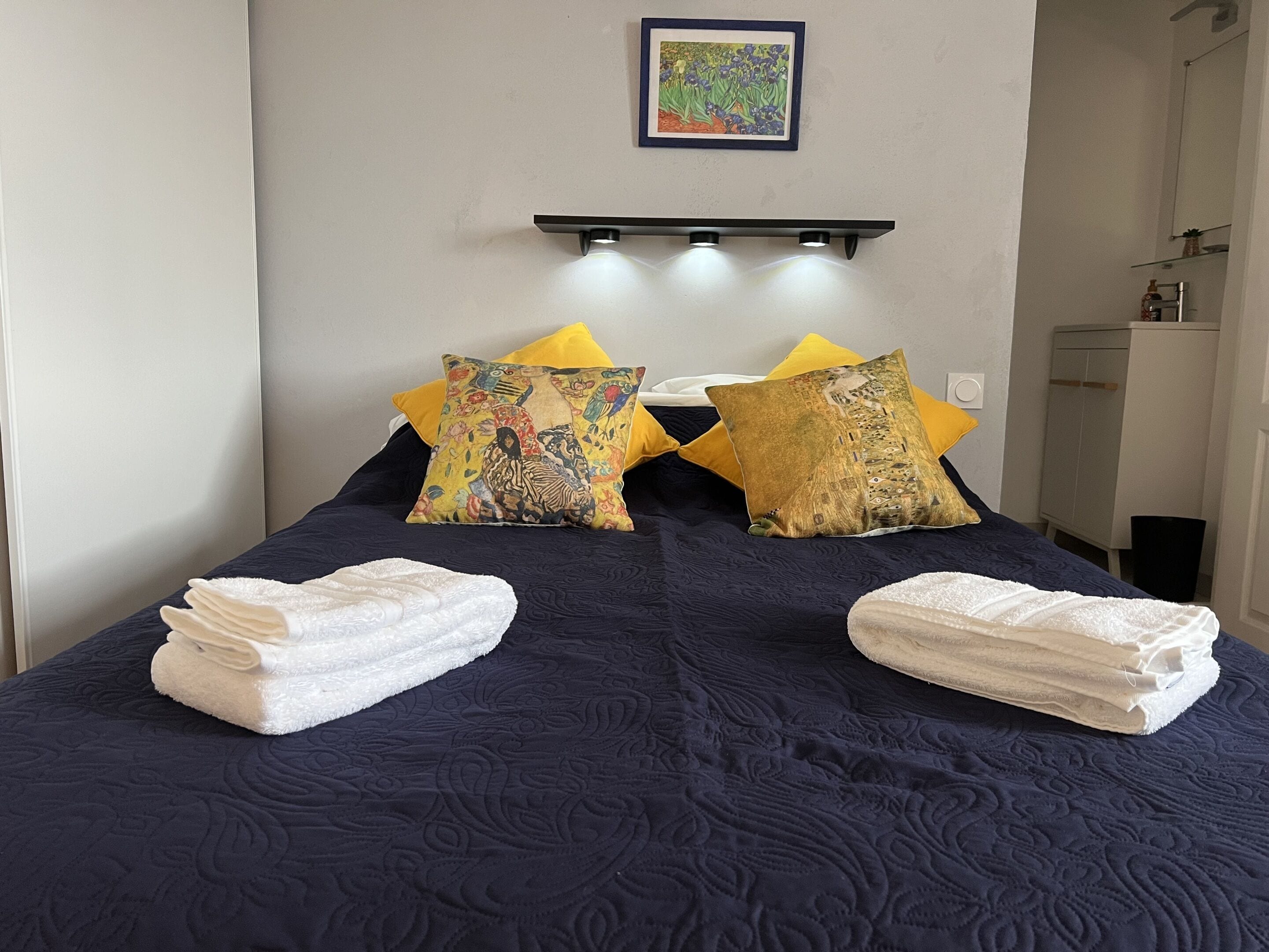 A neatly made bed with two decorative pillows, a bedside table, and a wall-mounted shelf with a framed picture.