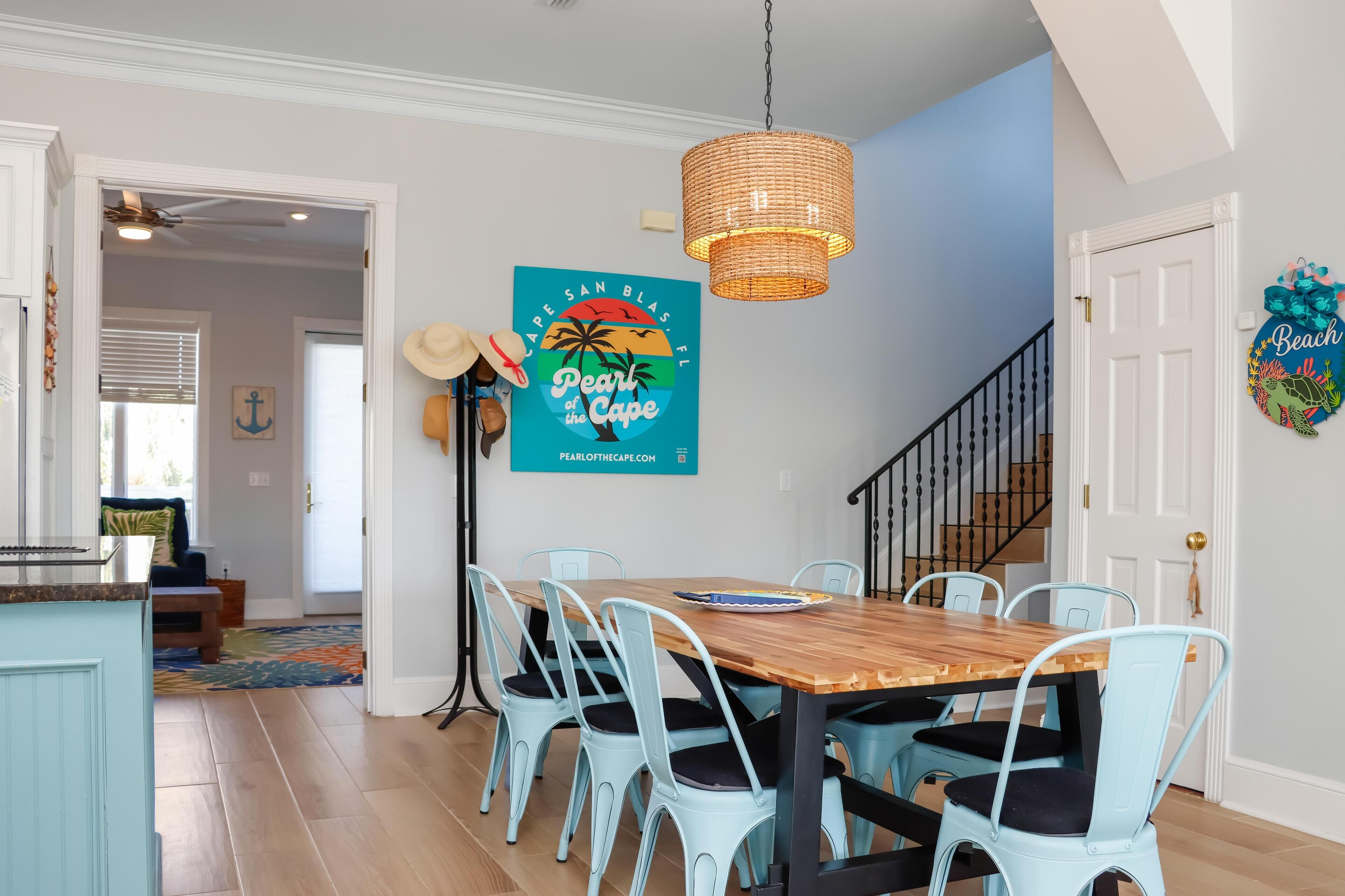 A dining area with a wooden table and light blue chairs, a hanging wicker chandelier, and a wall sign reading "Pearl of the Cape".