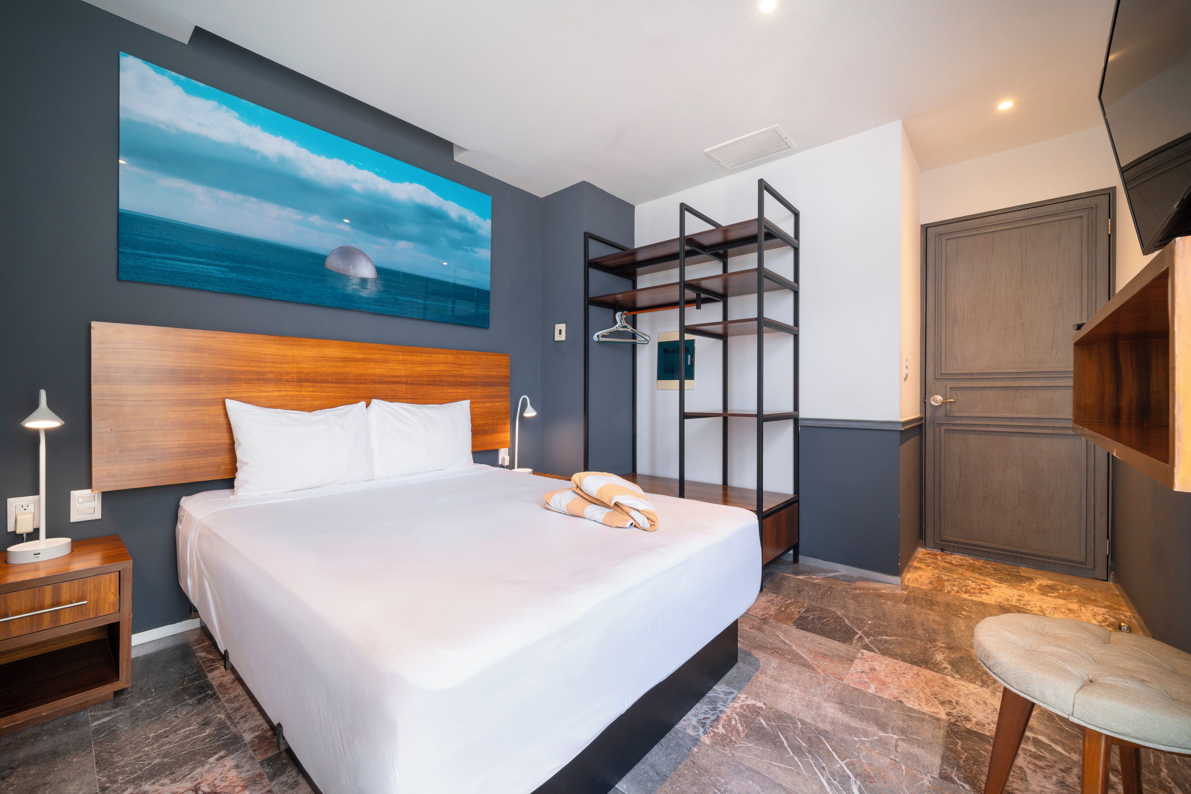 A hotel room with a large bed, a wooden headboard, a painting of the ocean, a built-in shelf, and a wardrobe.