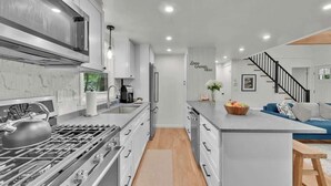 A modern kitchen with stainless steel appliances, a central island, and a staircase leading to a living area with a sofa and decorative pillows.