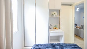 A bedroom with a bed, a wardrobe, a desk with a lamp, and a small shelf with decorative items.