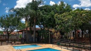 A swimming pool with a tiled deck, surrounded by lush greenery and a fenced area.