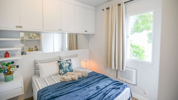 A bedroom with a bed, a window with curtains, a radiator, and a shelf with decorative items.