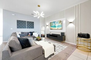 A modern living room with a grey sofa, a coffee table, a TV, and a wall-mounted speaker. - Private Pool, Free Parking, Solara Resort Home Near Disney, Accommodates 12 (Kissimmee)