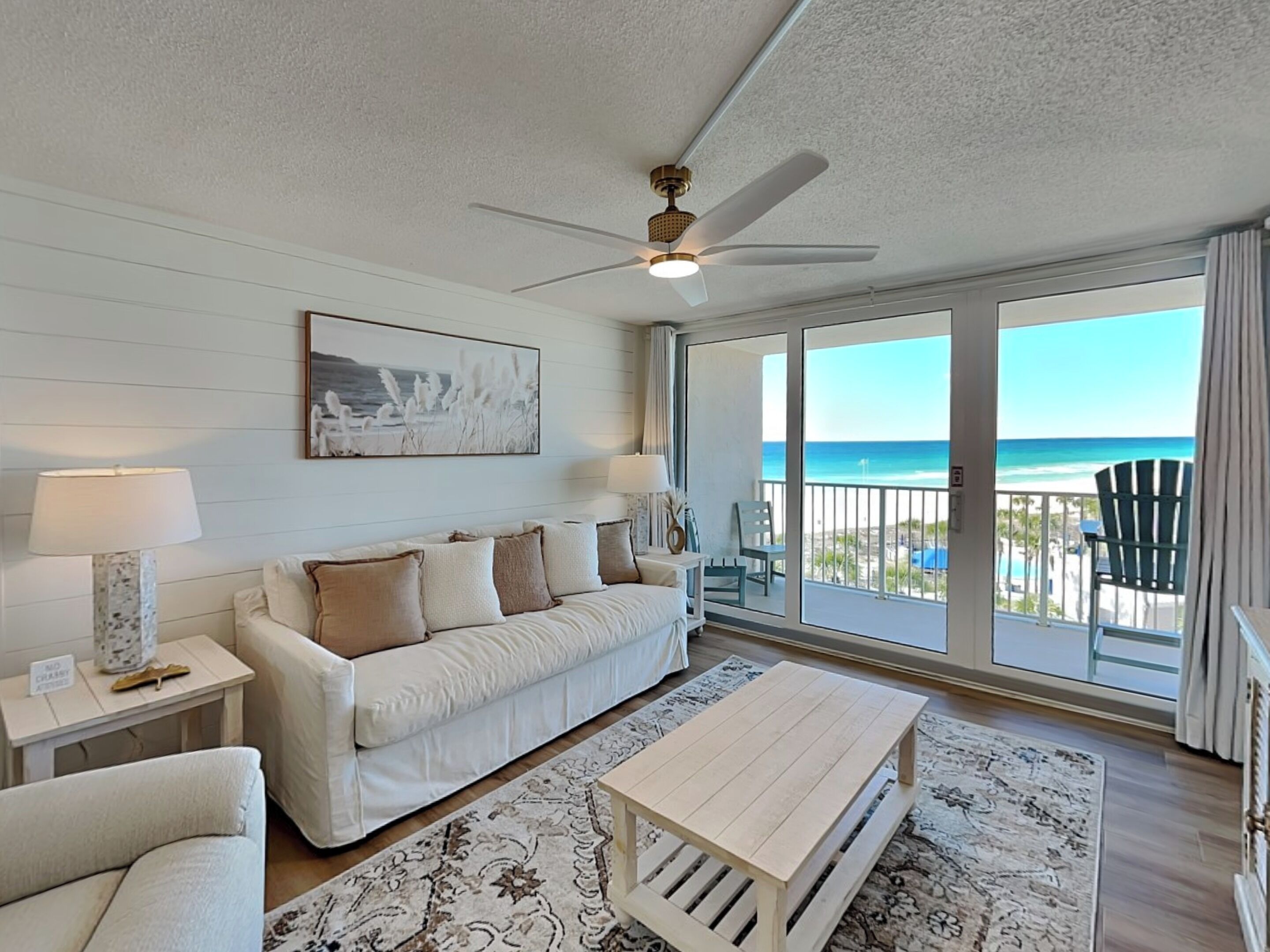 A spacious living room with a white sofa, a coffee table, and a ceiling fan. A large window offers a view of the beach.