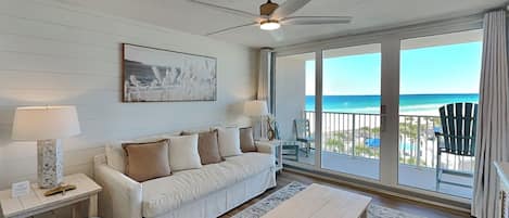 A spacious living room with a white sofa, a coffee table, and a ceiling fan. A large window offers a view of the beach.