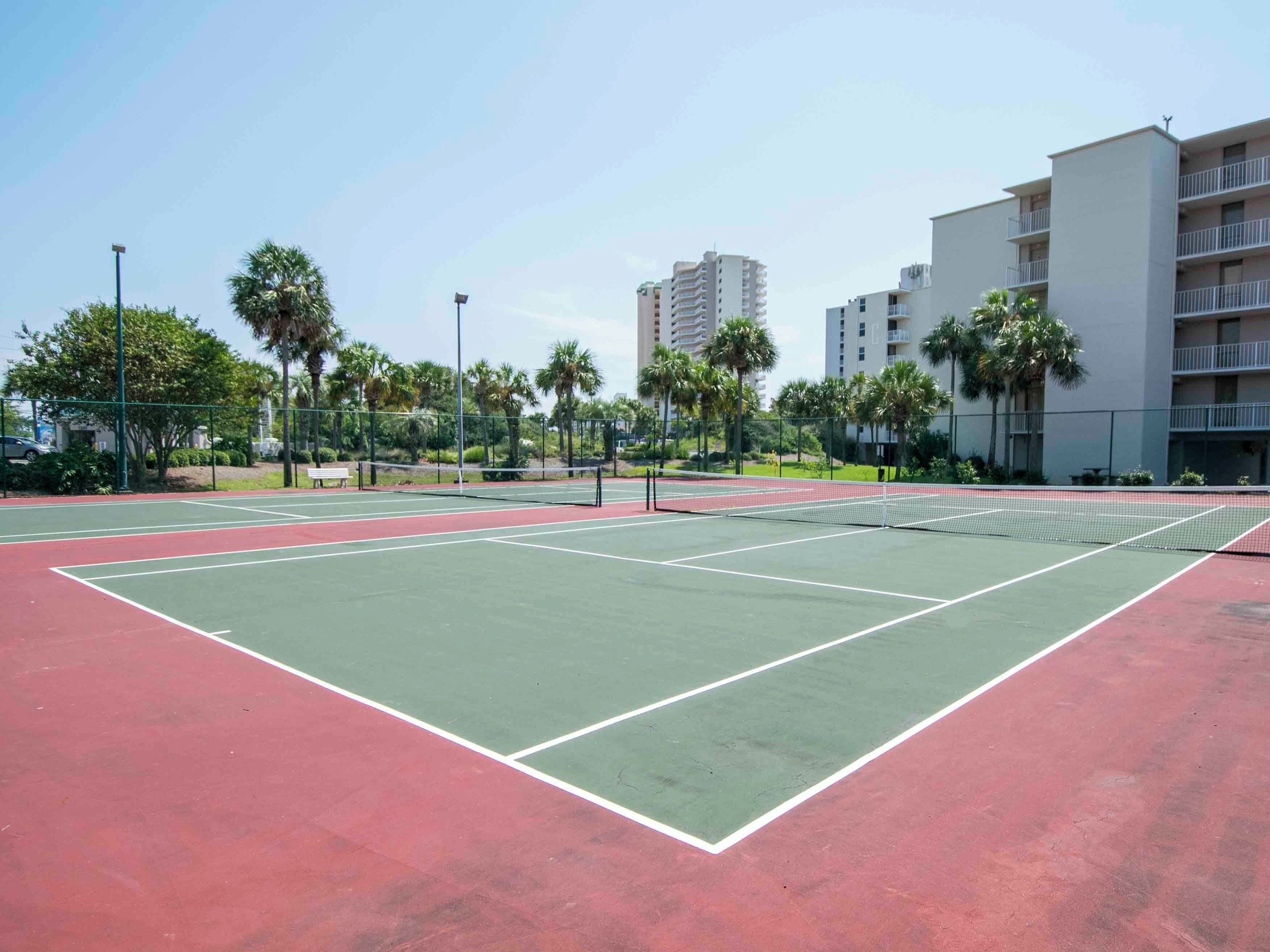 A tennis court with a net, surrounded by palm trees and a building in the background.