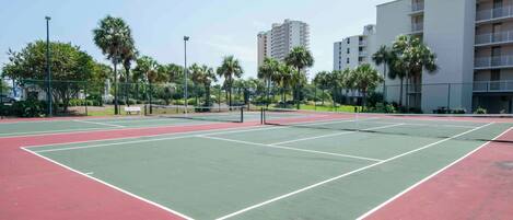 A tennis court with a net, surrounded by palm trees and a building in the background.