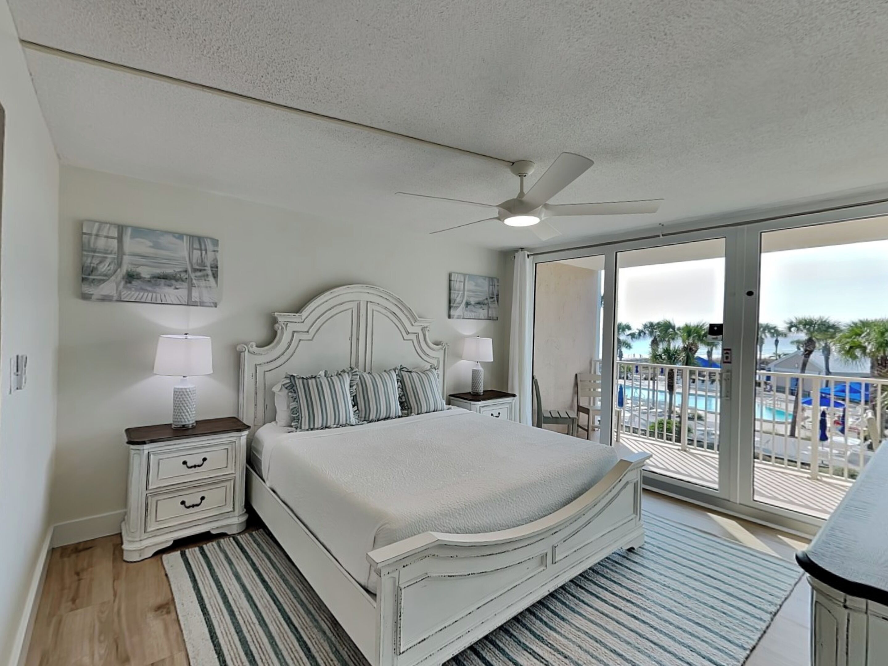 A bedroom with a bed, two nightstands, a ceiling fan, and a balcony with a pool view.