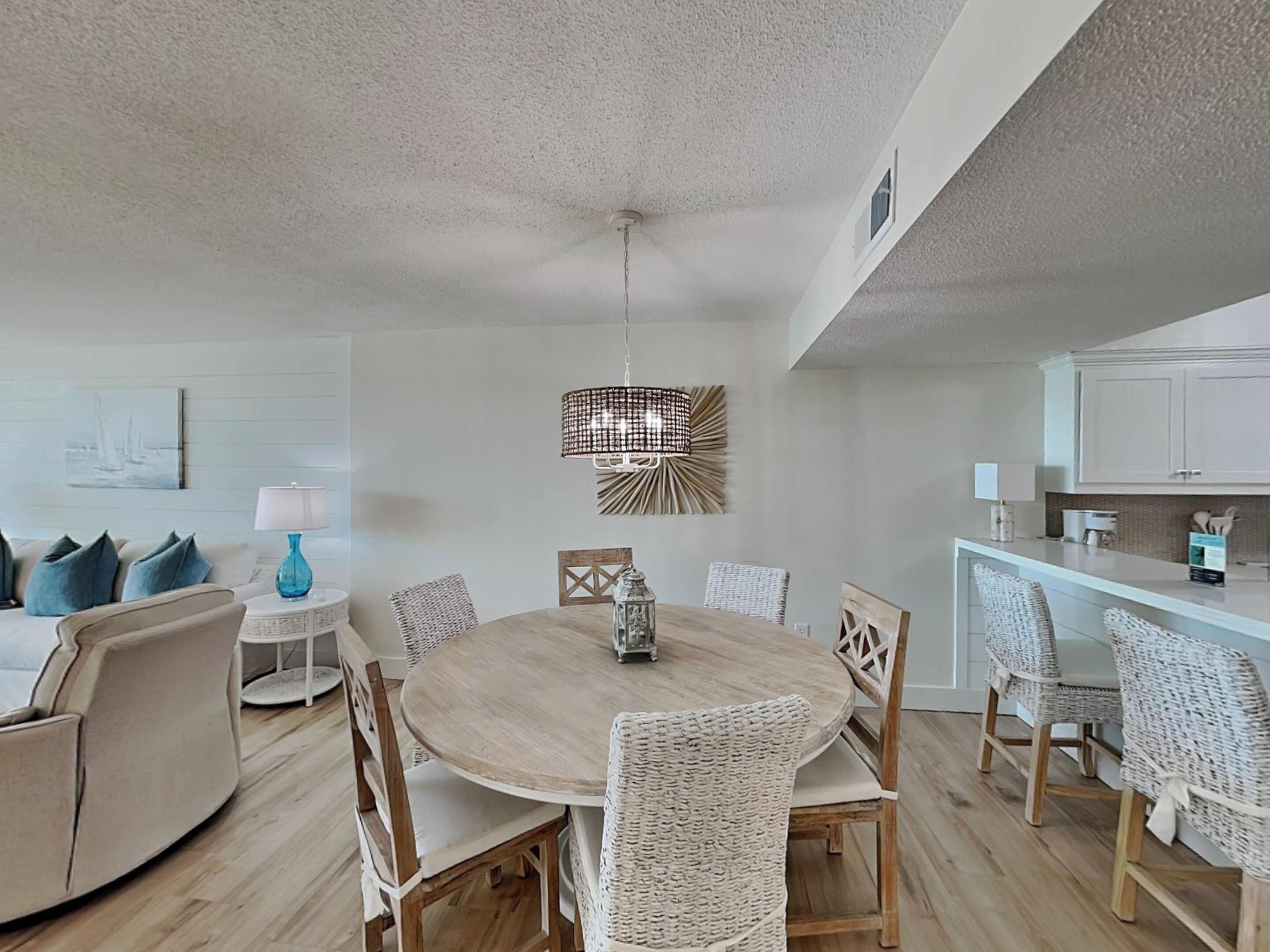 Un coin repas avec une table ronde, des chaises en osier, un luminaire suspendu et un espace cuisine doté de meubles blancs.