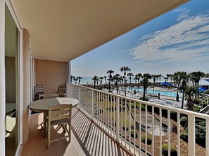 A balcony with a table and chairs, overlooking a pool area with palm trees and a beach.