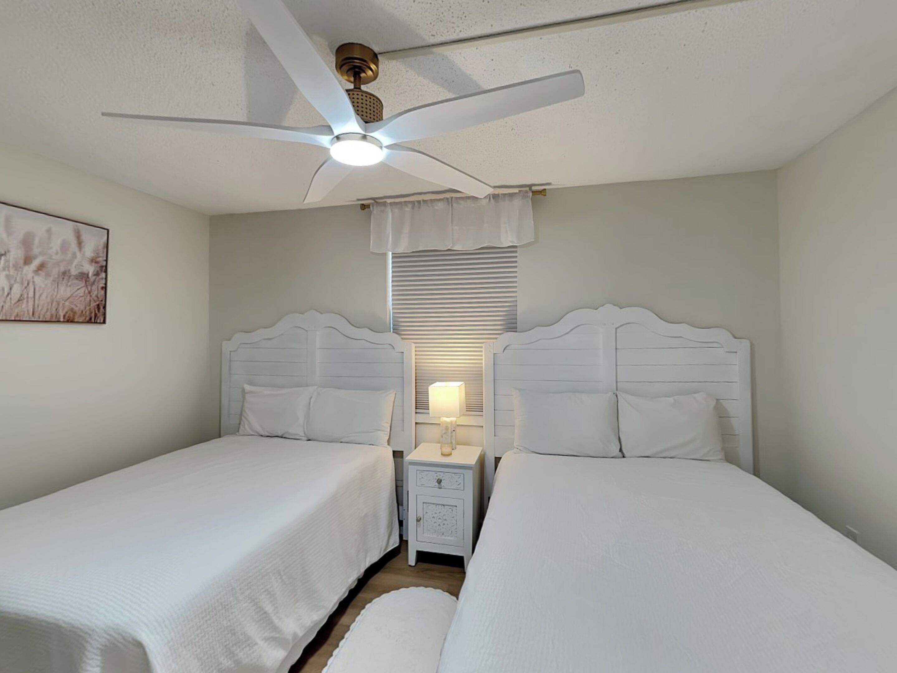 A twin bedroom with two beds, a ceiling fan, a nightstand with a lamp, and a window with blinds.