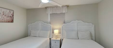 A twin bedroom with two beds, a ceiling fan, a nightstand with a lamp, and a window with blinds.