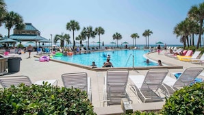 A pool area with lounge chairs, palm trees, and a building with a blue roof. - Dunes of Panama #c503 (Panama City Beach)