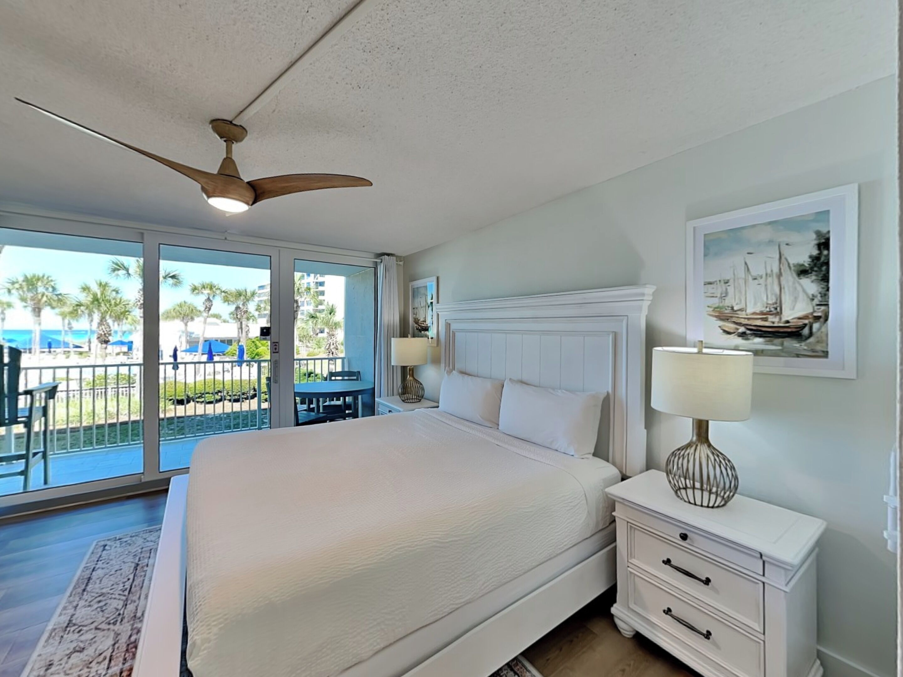 A hotel room with a large bed, a ceiling fan, a balcony with a view of palm trees and the beach, and a painting on the wall.