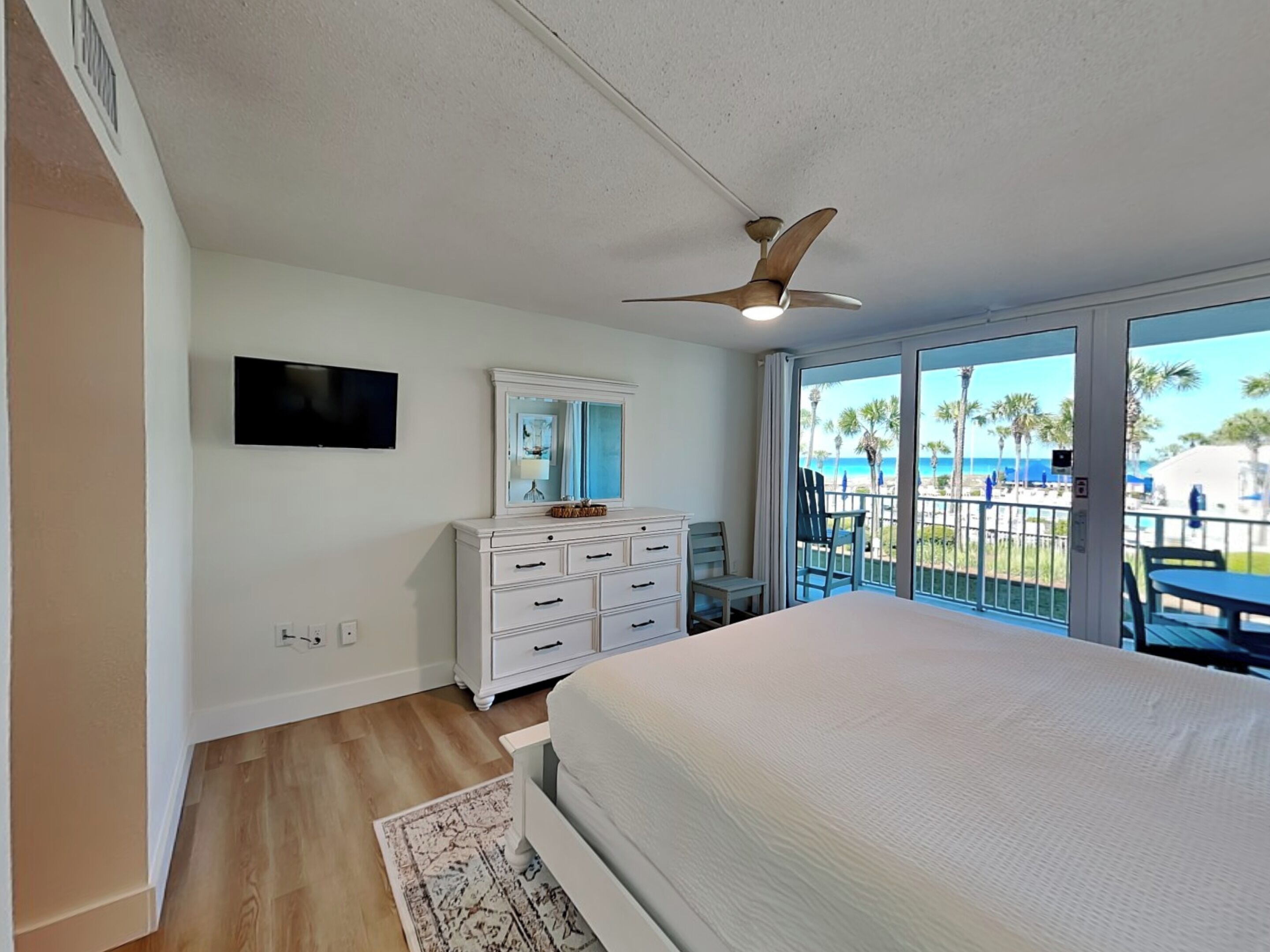Un dormitorio con cama, cómoda, ventilador de techo, televisor y balcón con vista a la playa.