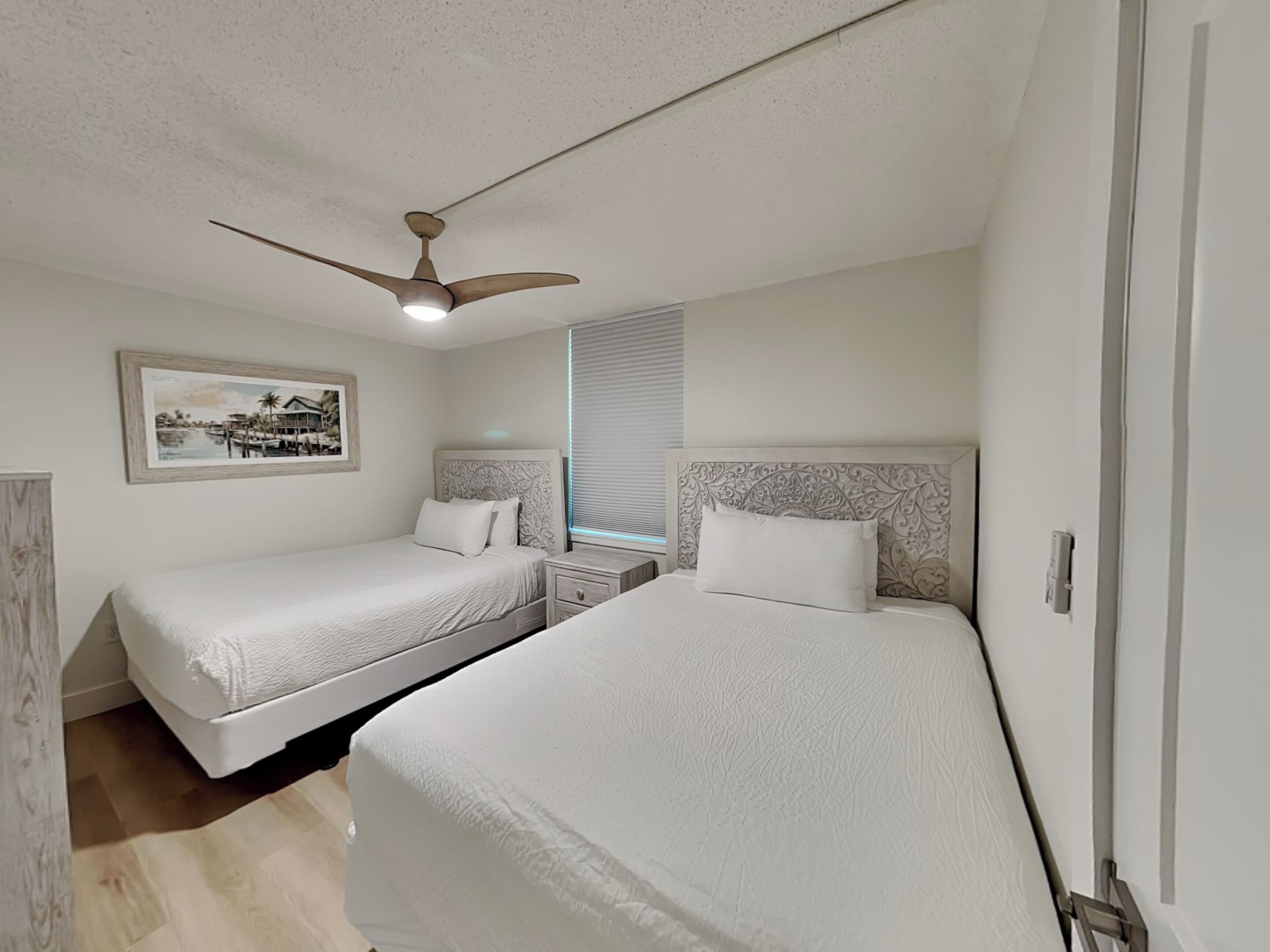 A hotel room with two beds, a ceiling fan, a nightstand, a framed picture, and a window with blinds.
