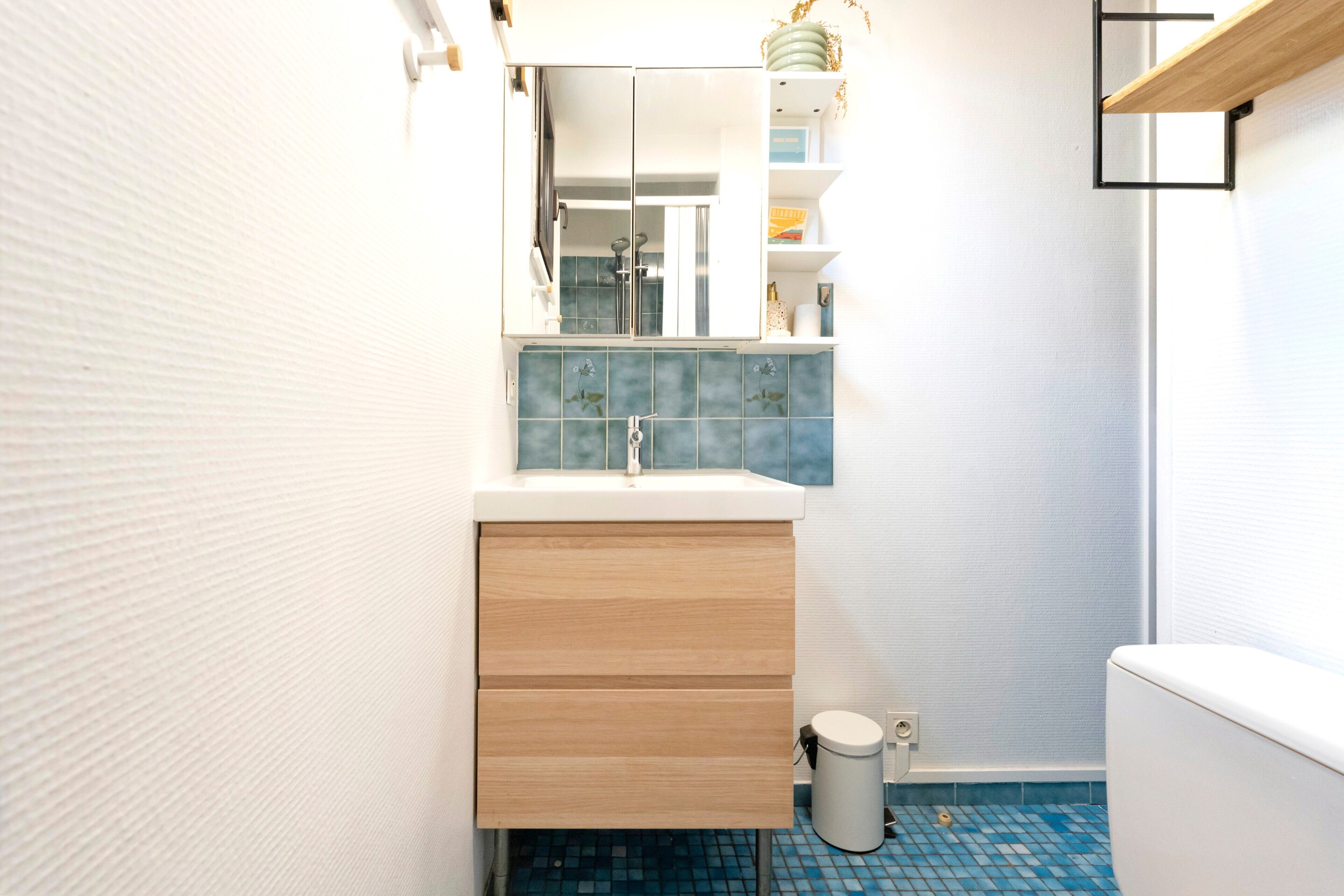 Une salle de bain avec un lavabo blanc, un miroir, un meuble en bois et une poubelle.
