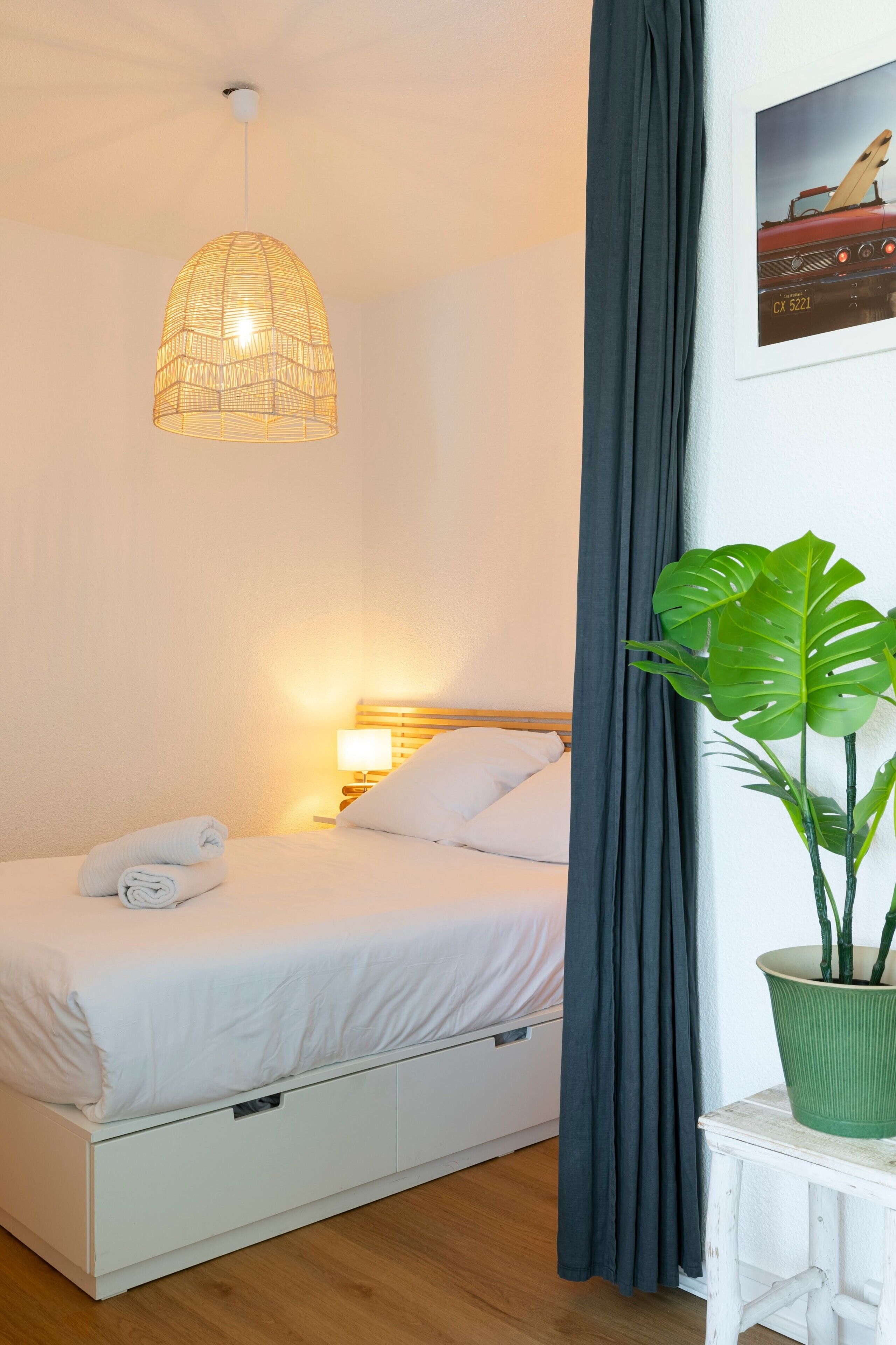 Een bed met wit beddengoed, een hanglamp, een donker gordijn en een plant in pot.