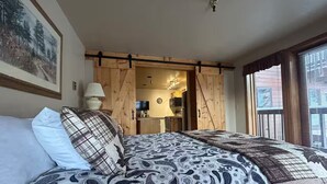Un dormitorio con cama, almohadas, una puerta de madera, televisor y ventana.