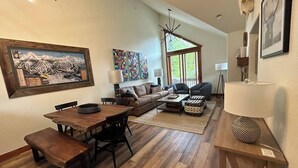 Un salon spacieux avec une table à manger en bois, un canapé et une table basse. Une grande fenêtre laisse entrer la lumière naturelle et offre une vue sur l’extérieur.