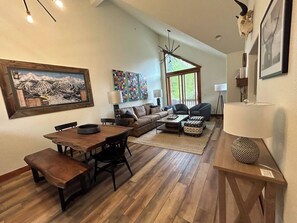 A spacious living room with a wooden dining table, a sofa, and a coffee table. There is a large window providing natural light and a view of the outdoors. - Ski~in/Ski~out~Saddlewood~3 Bd/3.5 Ba-Hot tub (Breckenridge)