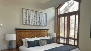 Une chambre avec un grand lit, une tête de lit en bois, une table de chevet avec une lampe, une fenêtre avec des stores et une œuvre d’art encadrée accrochée au mur.