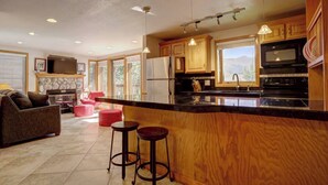 A modern kitchen with a large island, wooden cabinets, and a stone fireplace. There are two red bar stools and a sofa in the adjacent living area. - Ski~in~Ski~out~Tyra Aspen~2 Bd/2 Ba~Hot Tubs! (Breckenridge)
