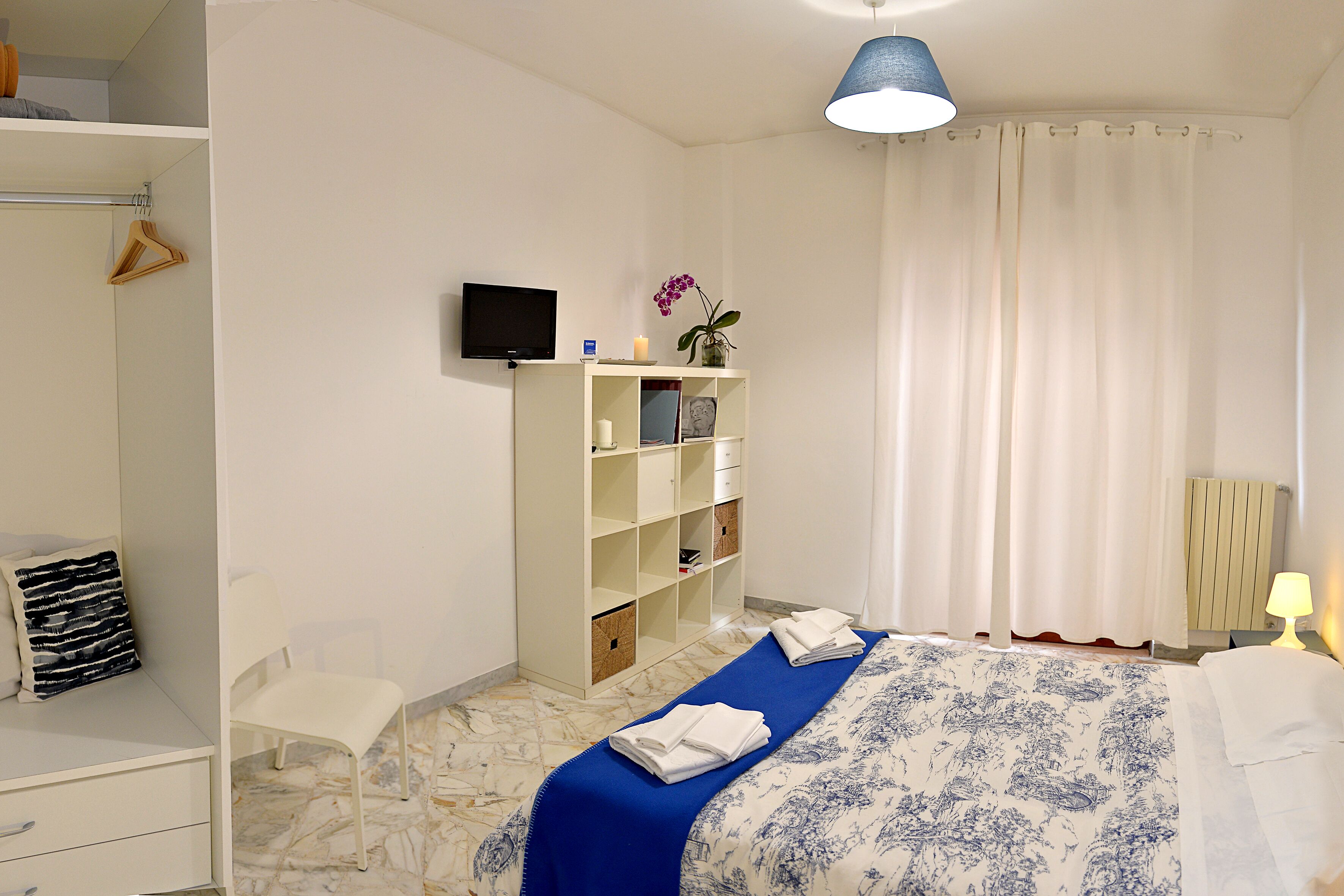 A bedroom with a bed, a wardrobe, a chair, a TV, and a bedside table with a lamp.