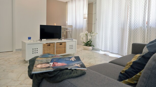 Ein modernes Wohnzimmer mit einem Flachbildfernseher auf einer weißen Konsole, einer Couch, einem Couchtisch mit Zeitschriften und einer Topfpflanze Orchidee.