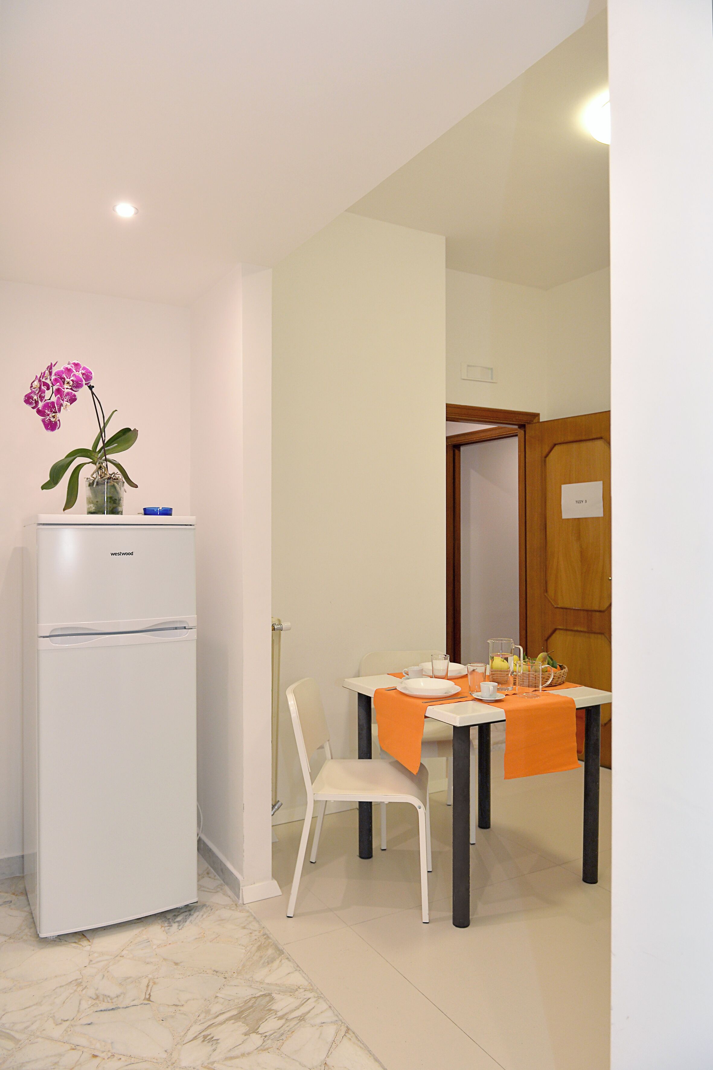 A compact room with a white refrigerator, a dining table with orange tablecloths, a white chair, and a small potted orchid.
