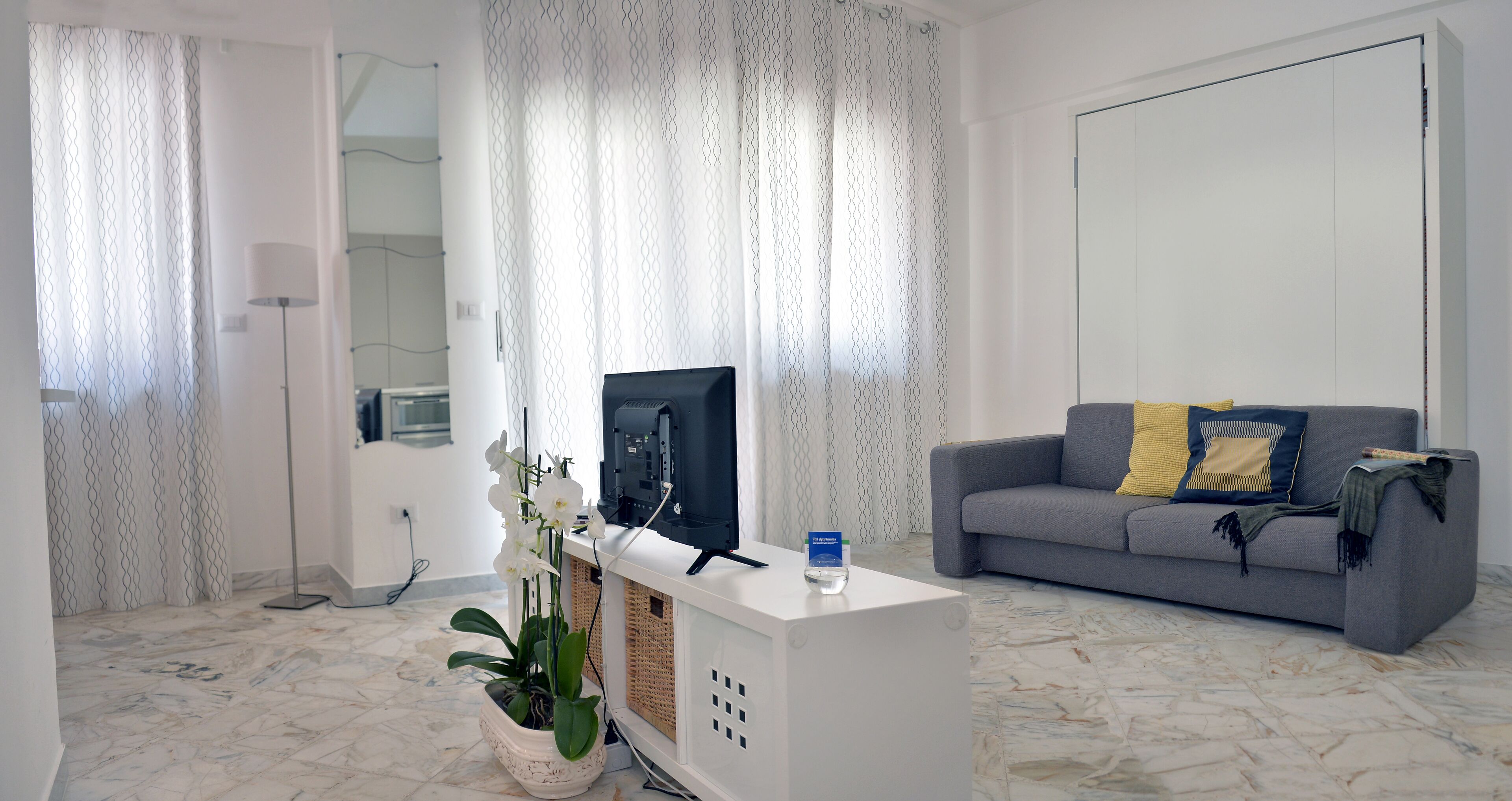 A living room with a grey sofa, a white entertainment unit, a potted orchid, and a television.