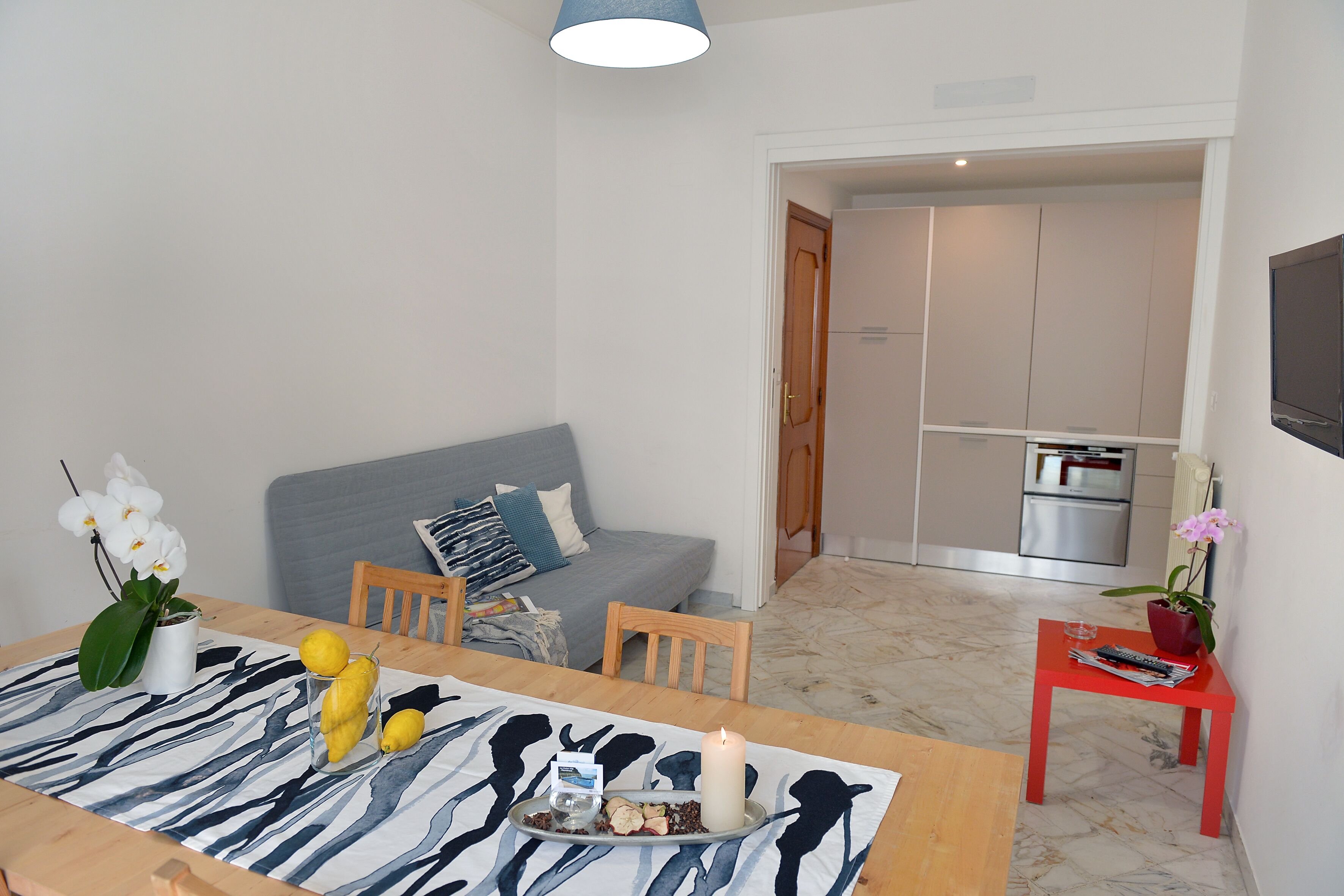 A dining area with a table set for two, a sofa, and a kitchen in the background.