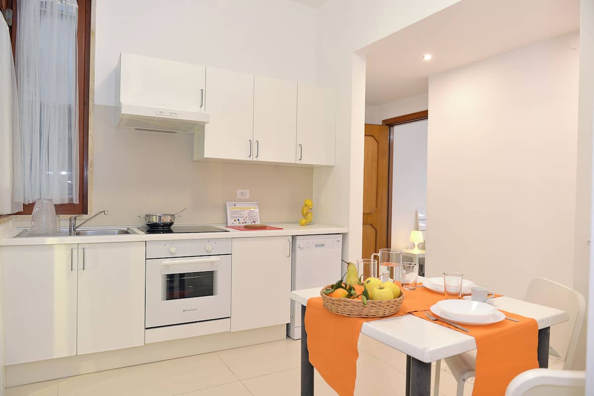 Une cuisine moderne avec des armoires blanches, un four encastré et un lave-vaisselle. On y trouve une table à manger recouverte d’une nappe orange, ainsi que des assiettes et des verres.
