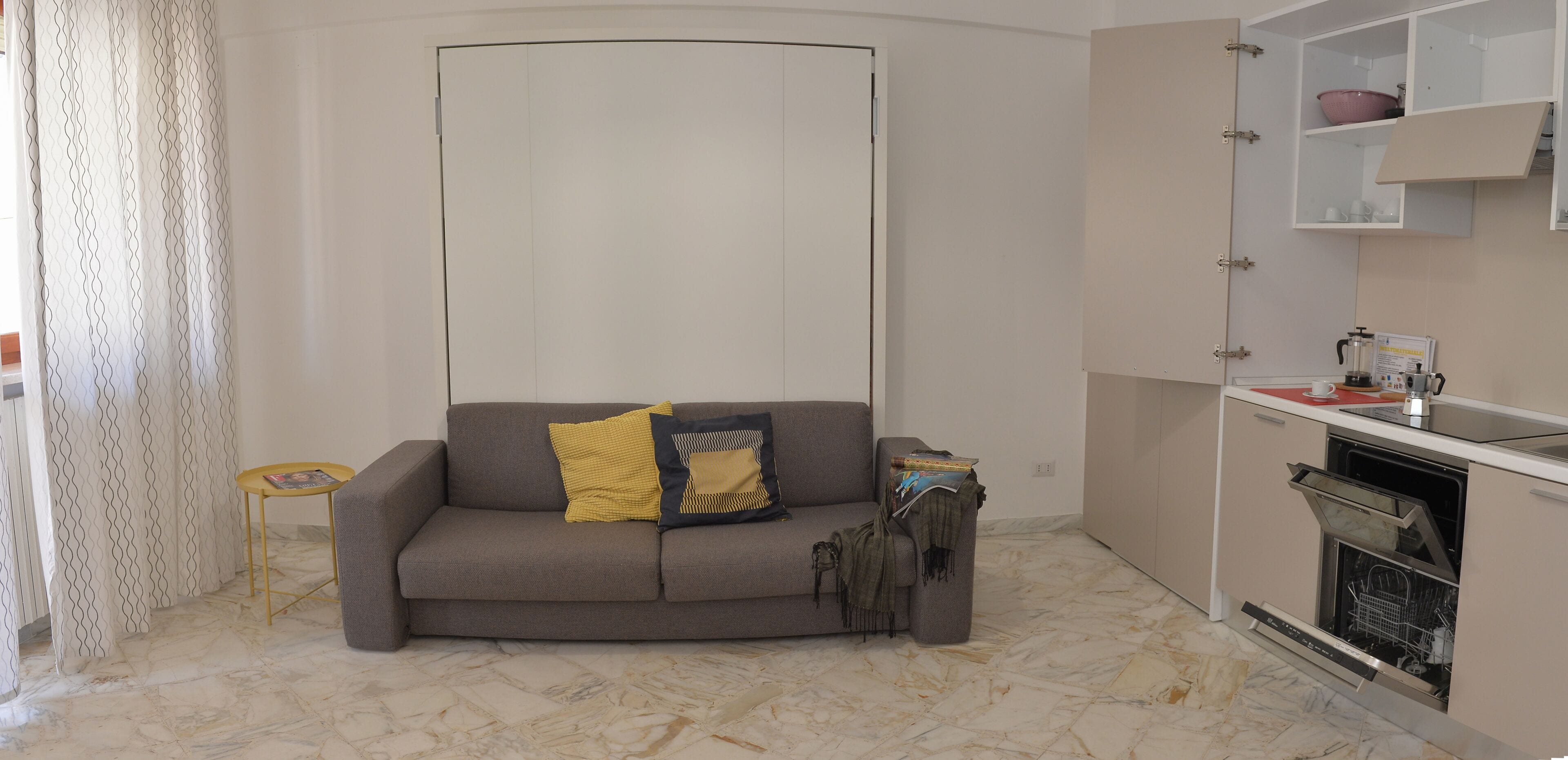 A compact living space with a sofa, a small table, a kitchen area with a dishwasher, and a patterned curtain.