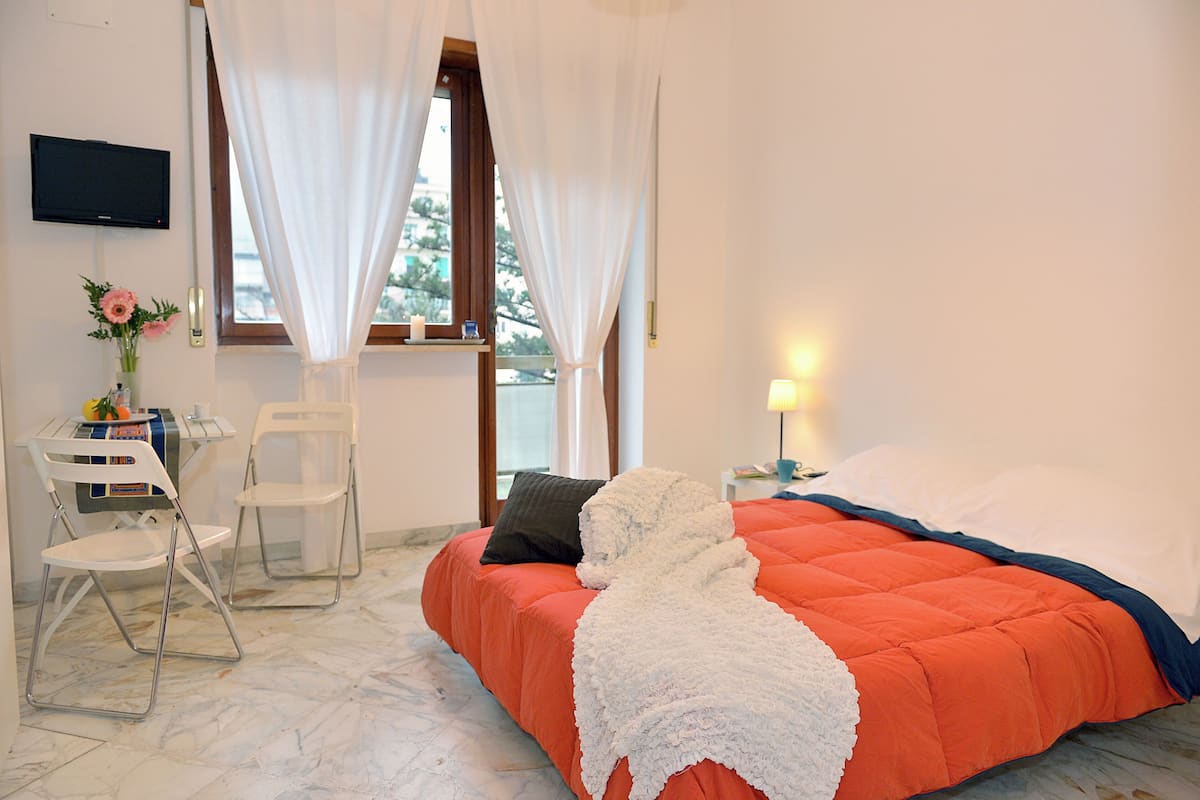 Une chambre à coucher avec un lit, un bureau avec des chaises, un téléviseur fixé au mur et une fenêtre avec des rideaux.